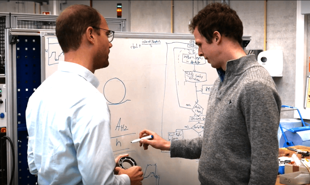 Ein Photo auf dem ein HCP Sense Experte einem Lager, insbesondere ein Wälzlager, in der Hand hält und der andere Experte Formeln auf einer weißen Tafel aufschreibt