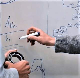 Eine Nahaufnahme auf der ein HCP Sense Experte einem Lager, insbesondere ein Wälzlager, in der Hand hält und der andere Experte Formeln auf einer weißen Tafel aufschreibt